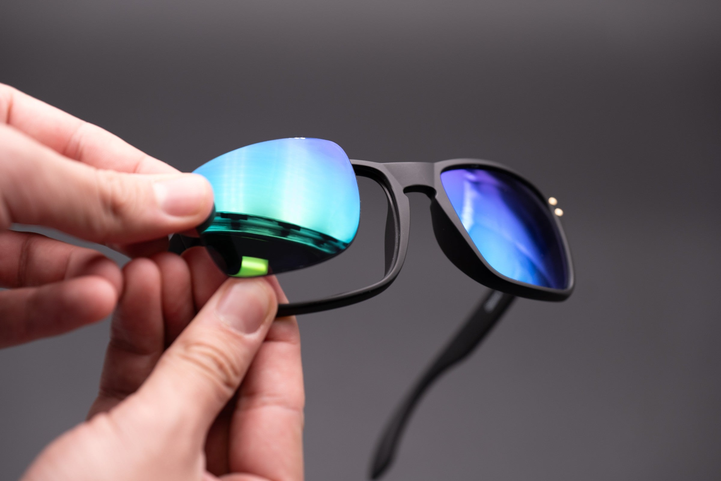 Hands snapping a mirrored teal replacement lens into a black sunglasses frame